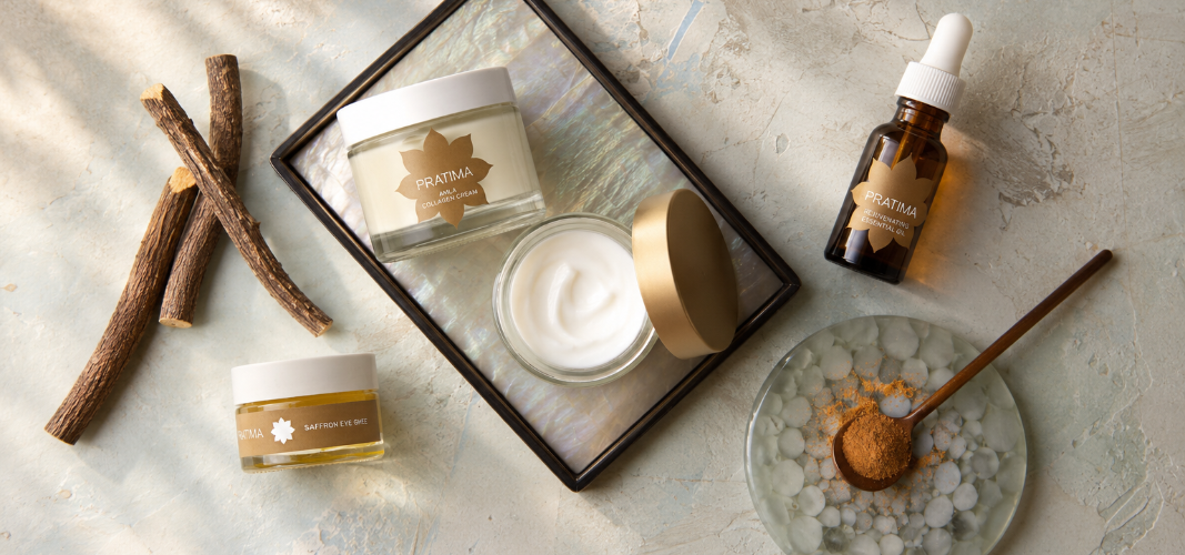 A styled flat-lay of skincare products on a textured light gray surface with dappled sunlight. The products include a jar of white cream in a glass jar with a gold lid, a small amber dropper bottle, two small cream jars, and a bowl with a wooden spoon containing a brown powder.