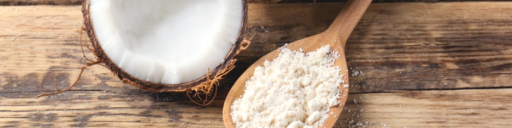 Coconut powder in a wooden spoon with a whole coconut and a halved coconut on a rustic wooden table.