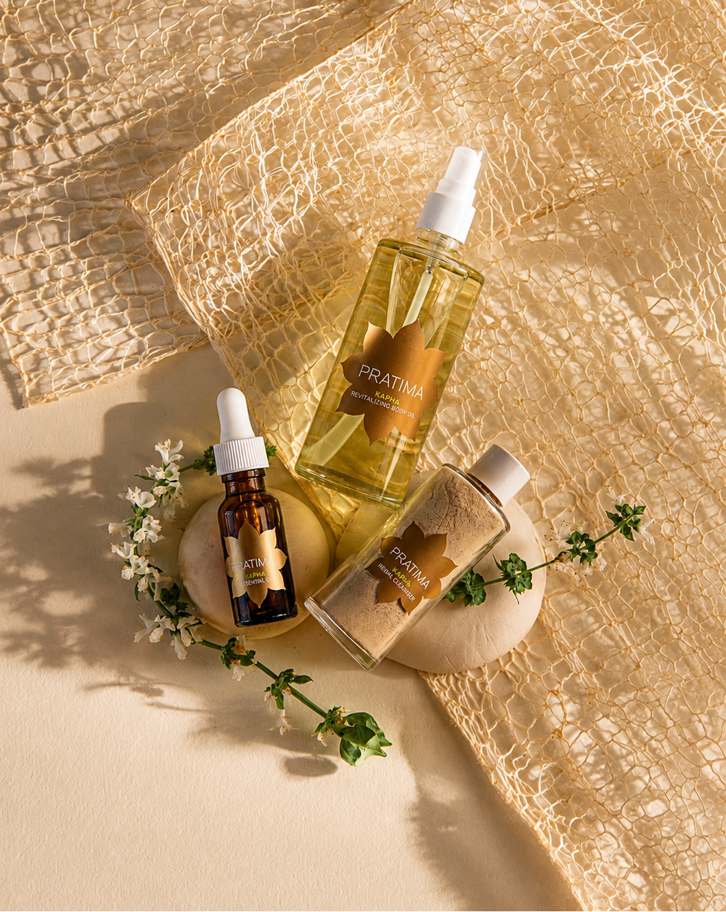 Three skincare products from the brand PRATIMA arranged on a textured beige surface with a gold mesh fabric backdrop. The set includes a large clear spray bottle filled with golden oil, a small amber dropper bottle, and a small jar of clay powder, styled with wooden blocks, white flowers, and green sprigs.