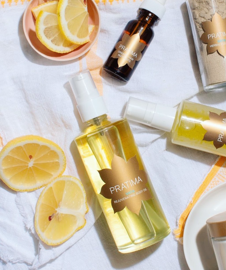 A styled flat-lay of PRATIMA Beeswax Hair Oil bottles on a white fabric background, surrounded by fresh lemon slices and a small dish of honey.